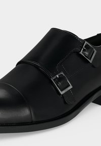Chaussure double boucles à brides pour homme en cuir noir avec boucles argentées et design à bout rapporté, vue en gros plan incliné.