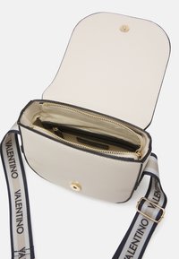 Borsa a tracolla beige con hardware dorato, bordo nero e tracolla regolabile a righe con il marchio 'Valentino'. Si apre su un interno foderato con tasche.