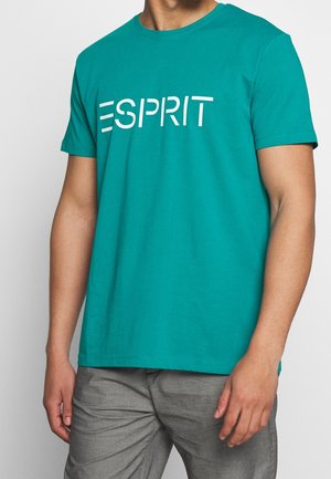 T-shirt en coton bleu sarcelle avec logo blanc "ESPRIT", col rond, manches courtes, texture lisse et coupe décontractée. Porté avec un pantalon gris clair.