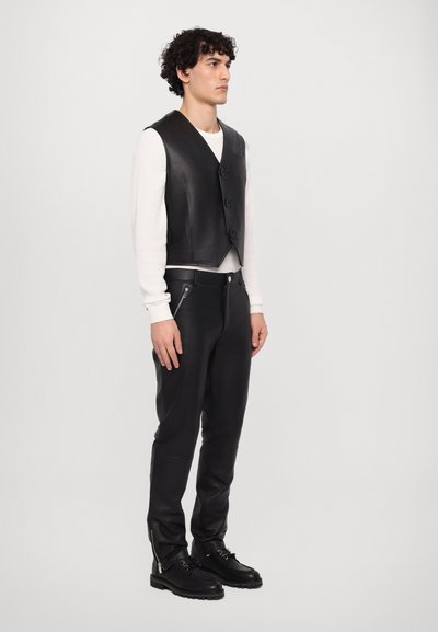 Gilet en cuir noir avec trois boutons, porté sur une chemise blanche à manches longues. Assorti à un pantalon noir ajusté et des bottes noires robustes.