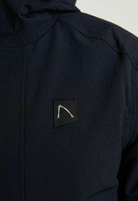 Veste bleu marine foncé avec un petit patch noir présentant un design abstrait blanc minimaliste sur la partie supérieure de la poitrine.