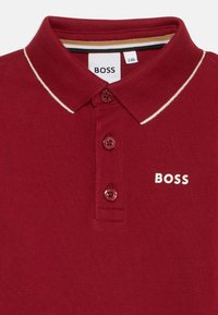 Burgundi színű polo póló, texturált anyagból, két gombos gallérral, krém színű díszítéssel a galléron, és fehér "BOSS" logóval.