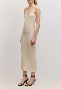 Beige strukturiertes Strick-Maxikleid mit dünnen Trägern, das eine figurbetonte Silhouette und Seitenschlitze aufweist. Kombiniert mit schwarzen, dünngestreiften, hochhackigen Sandalen.
