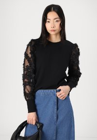 Schwarzer Pullover mit strukturierten schwarzen floralen Verzierungen an durchsichtigen Ärmeln, kombiniert mit einem Jeansrock und einer schwarzen Tasche.