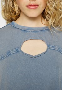 Ljusblå bomullstop med rundad halsringning och ett stort, oregelbundet utskärning nedanför. Har fransade kanter och en mjuk textur.