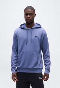 Jeune homme portant un sweat à capuche Nike bleu et un pantalon Nike Pro noir, se tenant devant un fond uni gris clair.