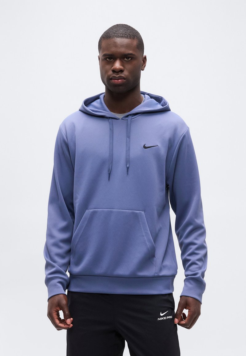 Jeune homme portant un sweat à capuche Nike bleu et un pantalon Nike Pro noir, se tenant devant un fond uni gris clair.