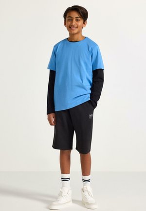 Teini-ikäinen poika pukeutuneena siniseen ja mustaan pitkähihaiseen sekä lyhythihaiseen paitaan, mustiin shortseihin, valkoisiin raidallisiin sukkiin ja valkoisiin tennareihin, seisoo ja hymyilee.