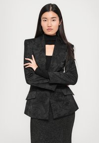 Black blazer with textured floral pattern, peaked lapels, and two front pockets. Paired with a fitted black top and textured gray skirt.