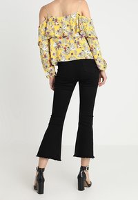 Top giallo a fiori con spalle scoperte e balze abbinato a jeans neri a zampa d'elefante. I jeans hanno orli grezzi e sono indossati con sandali neri con tacco.