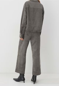 Persona vestita con una giacca in corduroy grigia e pantaloni a gamba larga, con stivali neri con tacco alla caviglia, in piedi su uno sfondo neutro.