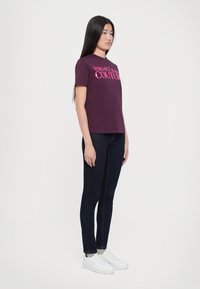 Bordo marškinėliai su užrašu "VERSACE LA COUTURE" rožine spalva, derinami su tamsiai mėlynomis aptemptomis džinsomis ir baltomis sportbačiais. Lygus audinys ir klasikėmis kirpimo.