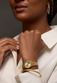 Montre-bracelet en or avec un cadran rond et une lunette texturée, associée à un bracelet en or torsadé ; le modèle porte une chemise blanche à col.