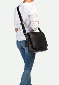 Borsa a traverso in pelle nera con chiusura a patta, tracolla regolabile e etichetta per bagagli. Texture liscia e design minimalista.