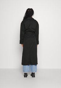 Svart trenchcoat med belte i midjen, lange ermer og en subtil glans. Båret over lyseblå vide jeans og svarte støvler.
