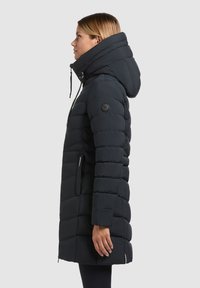 Zwarte pufferjas met verticaal gewatteerd patroon, lange lengte, hoge kraag en capuchon met trekkoord; voorzien van een logo-badge op de mouw.