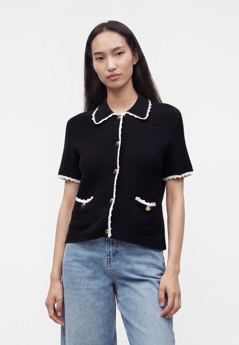 Femme portant un cardigan en maille noir à manches courtes avec finition blanche et boutons dorés, associé à un jean bleu clair, debout devant un fond uni.