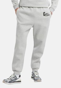 Person iført lysegrå sweats med "Carlo Colucci" logo på låret og mønstrede sort-hvide sneakers, hænderne i lommerne.