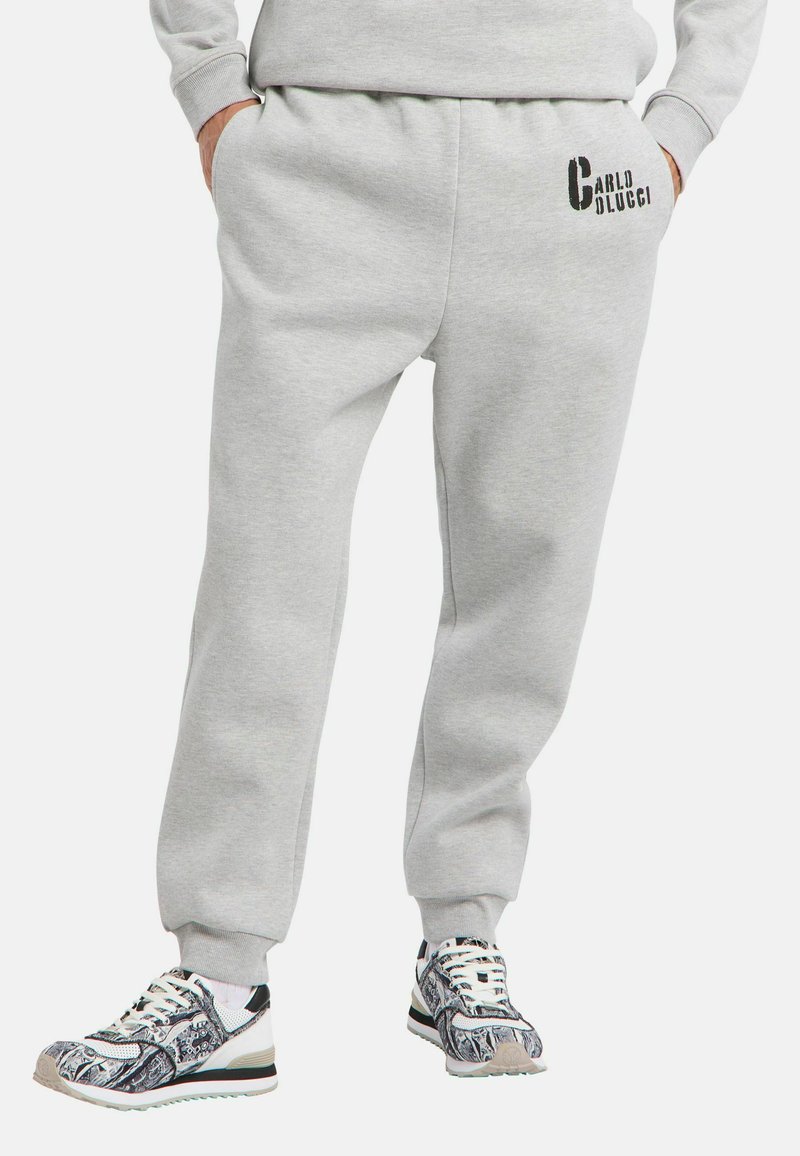 Person iført lysegrå sweats med "Carlo Colucci" logo på låret og mønstrede sort-hvide sneakers, hænderne i lommerne.