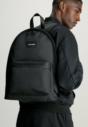 Schwarzer Rucksack aus strukturiertem Stoff, mit einer vorderen Reißverschlusstasche, Logo-Patch und gepolsterten Schultergurten sowie einem praktischen Tragegriff.