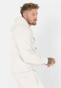 Hvit hettegenser med kengurulomme og ribbestrikkede mansjetter. Har snor i hetta og logo trykt på fronten. Myk, strukturert stoff.