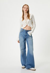 Weißes, strukturiertes Crop-Top mit Fransen an den Rändern und gebundenem Frontteil. Hellblaue Schlagjeans mit hoher Taille. Modell trägt weiße Sneakers.