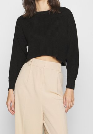 Femme portant un pull court en tricot noir à manches longues et un pantalon beige taille haute à jambes larges sur un fond uni.
