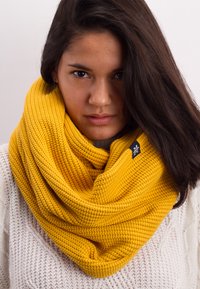 Knit yellow infinity scarf featuring a textured pattern and a small black label. Worn over a light-colored sweater.