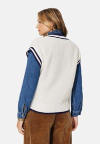 Sweater sans manches en tricot blanc avec des rayures bleu marine, superposé sur une chemise en denim bleue. Présente des détails de bord-côte à l'ourlet et aux poignets.