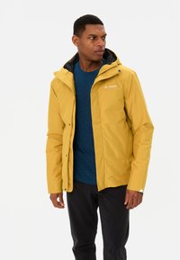 Gele waterdichte jas met een capuchon, met een zwarte binnenvoering, een rits aan de voorkant en een logo op de borst. Draagt over een blauw shirt.