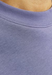 Nahaufnahme einer Person, die einen lavendelfarbenen, gerippten Rundhalsausschnitt-Pullover trägt, wobei die Stoffstruktur und die Details des Ausschnitts zu sehen sind.