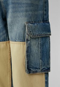 Jeans med en kontrasterande beige canvaspanel, utrustade med en stor cargoficka på sidan och synliga sömnadsdetaljer.