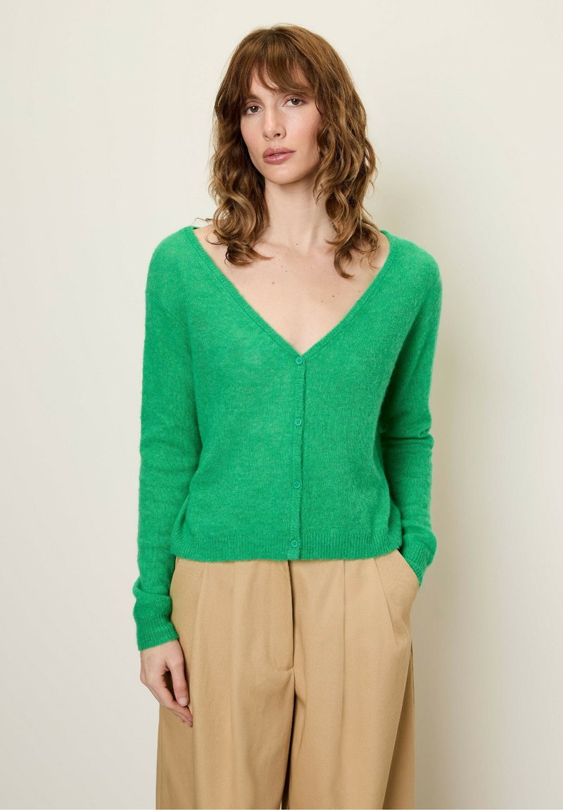 Groene, zachte, textuur cardigan met een diepe V-hals en knoopsluiting. Lange mouwen en een getailleerde taille, gecombineerd met lichtbeige broeken.