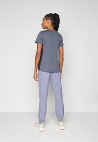 Grå sporty T-skjorte med korte ermer, kombinert med lyse lilla joggebukser. Joggebuksene har elastiske mansjetter; antrekket inkluderer lyse treningssko.