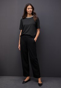 Top corto negro texturizado de manga corta con un ribete blanco, combinado con pantalones anchos negros y zapatos planos negros puntiagudos, frente a un fondo gris.