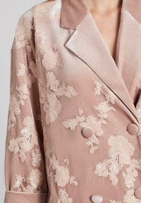Blazer de terciopelo suave en rosa empolvado, de doble botonadura, que presenta intrincados bordados florales y lentejuelas. Notables detalles de la textura del tejido y de los botones.
