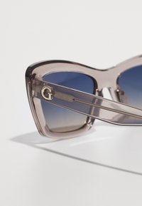 Angular, oversized sunglasses with a translucent mauve frame, gradient blue lenses, and gold 'G' logo on the temple.