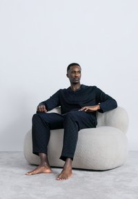 Férfi fekete pizsamában, mezítláb ülve egy világos bézs, lekerekített modern székben, egy egyszerű fehér fal előtt.