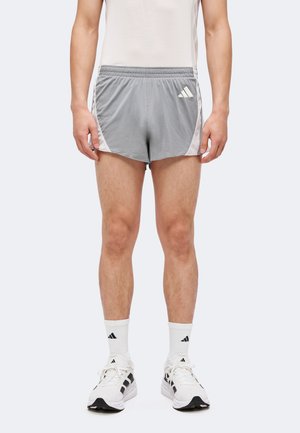 Homme portant un short de course gris Adidas, des chaussettes blanches, des baskets blanches Adidas, debout devant un fond uni.