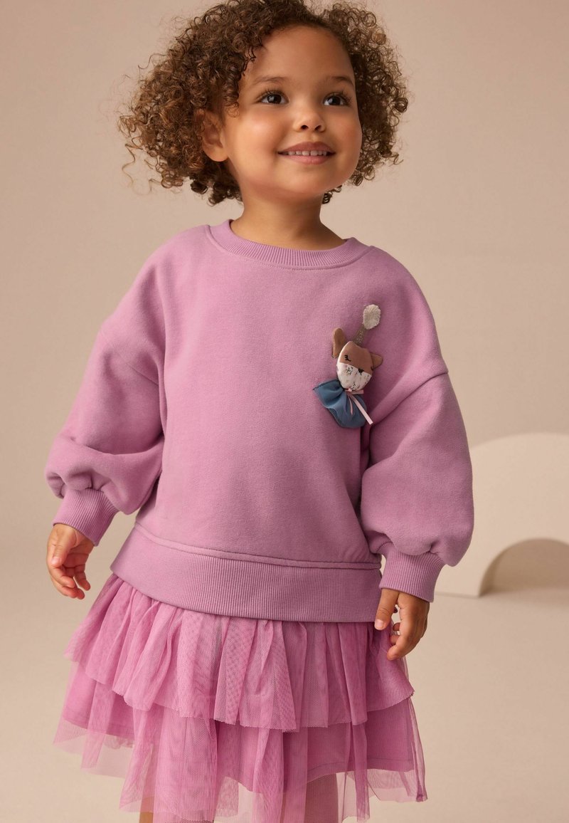 Jeune enfant aux cheveux bouclés portant un sweat-shirt rose avec une broche en forme de chat et une jupe en tulle rose à plusieurs couches, souriant et regardant légèrement vers le haut.