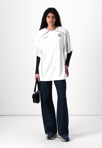 Hvit oversized jersey med kort erm, detaljert søm og en sirkulær logo. Kombinert med mørkeblå utsvungne jeans og en svart håndveske.