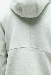 Πίσω μέρος ανοιχτόχρωμου γκρι φλις μπουφάν με κουκούλα και εμφανείς ραφές, με μικρό λευκό λογότυπο "MAMMUT" τοποθετημένο στο κέντρο κάτω από την κουκούλα.