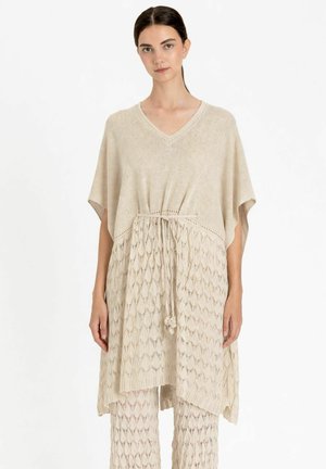 Donna che indossa una tunica beige in maglia con coulisse in vita e pantaloni coordinati in maglia a motivo, in piedi su sfondo bianco.