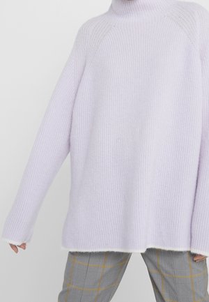 Pull côtelé lavande à manches longues porté avec un pantalon à carreaux gris présentant des rayures jaunes, montrant le torse et le haut des jambes.