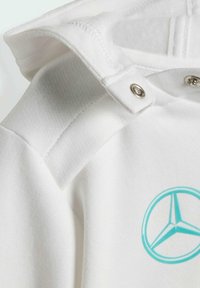 Weißer Hoodie mit einem türkisfarbenen Mercedes-Logo, metallischen Druckknöpfen und einem weichen, strukturierten Stoff am Kragen und den Schultern.