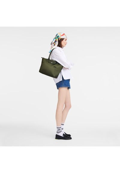 Olive green tote bag with black hardware, featuring a rounded shape and short handles. Model wears a white shirt, denim shorts, and loafers.