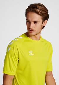 Heldere gele sportieve shirt met korte mouwen, gemaakt van gladde stof, met een wit logo en chevronpatronen op de schouders.
