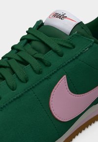 Baskets en daim vert avec un swoosh en cuir rose, des lacets verts texturés et une étiquette blanche avec le logo "Nike" rouge sur la languette.
