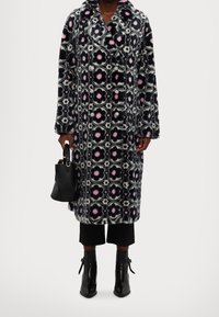 Cappotto multicolore con motivo floreale in tessuto morbido, caratterizzato da uno sfondo nero, accenti rosa e bianchi, e grandi bottoni. Indossato sopra pantaloni e stivali neri.