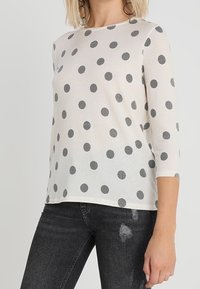 Weißes, semitransparentes Oberteil mit Dreiviertelärmeln, verziert mit großen schwarzen Punkten. Kombiniert mit ausgefransten schwarzen Jeans.